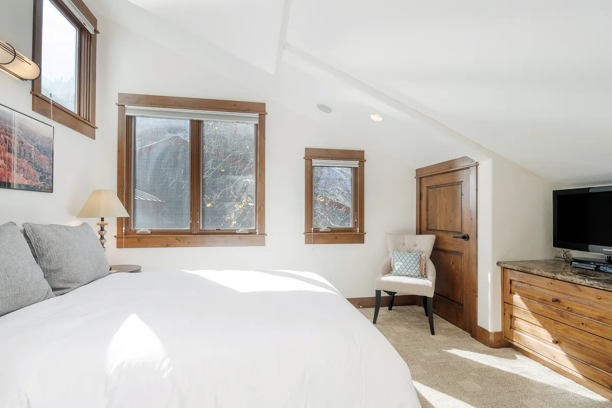 The image showcases a serene bedroom with a plush bed adorned in crisp white bedding, warm wooden accents, and large windows flooding the space with natural light. The atmosphere invites relaxation, with thoughtful touches that encourage guests to unwind, such as reading in bed or enjoying a quiet morning coffee while gazing at mountain scenery.
