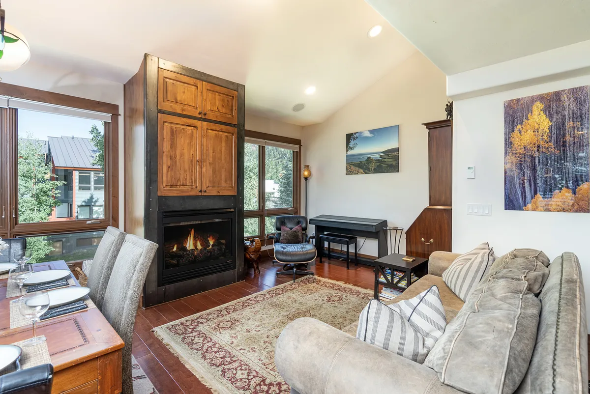 This image captures a warm and welcoming living room featuring a prominent gas fireplace, a dining table, and large windows that flood the room with natural light. Family members are gathered near the fireplace, while others relax and play music at a nearby piano, all set against the backdrop of the expansive mountain views outside.