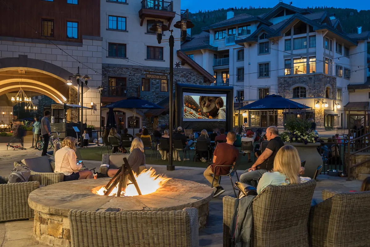 An inviting exterior scene featuring a cozy fire pit, an outdoor movie screen, and comfortable seating. The building’s architecture is visible, and people are gathered, enjoying the warmth and communal spirit under the evening sky.