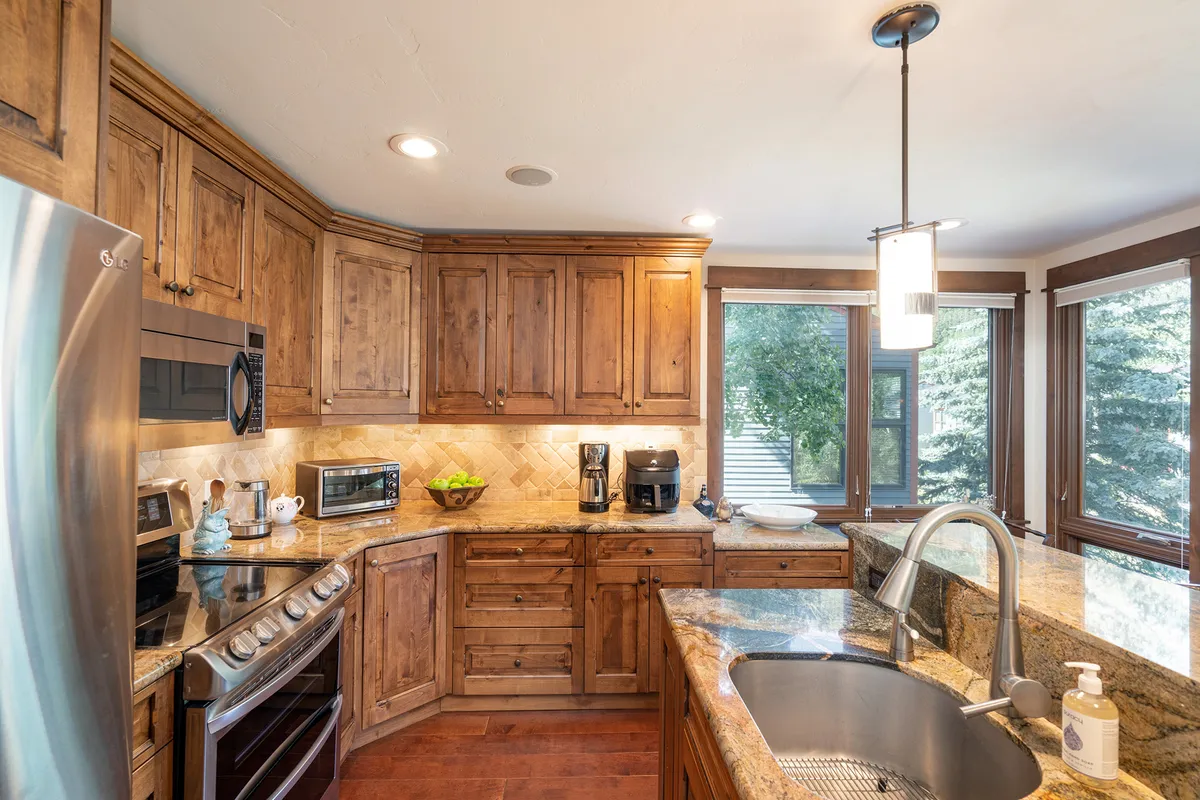 A warmly lit, inviting kitchen with granite countertops and wooden cabinets, featuring modern appliances and ample counter space. Large windows allow natural light to fill the space, and there are hints of family gatherings and cooking activities, creating a cozy, welcoming atmosphere.