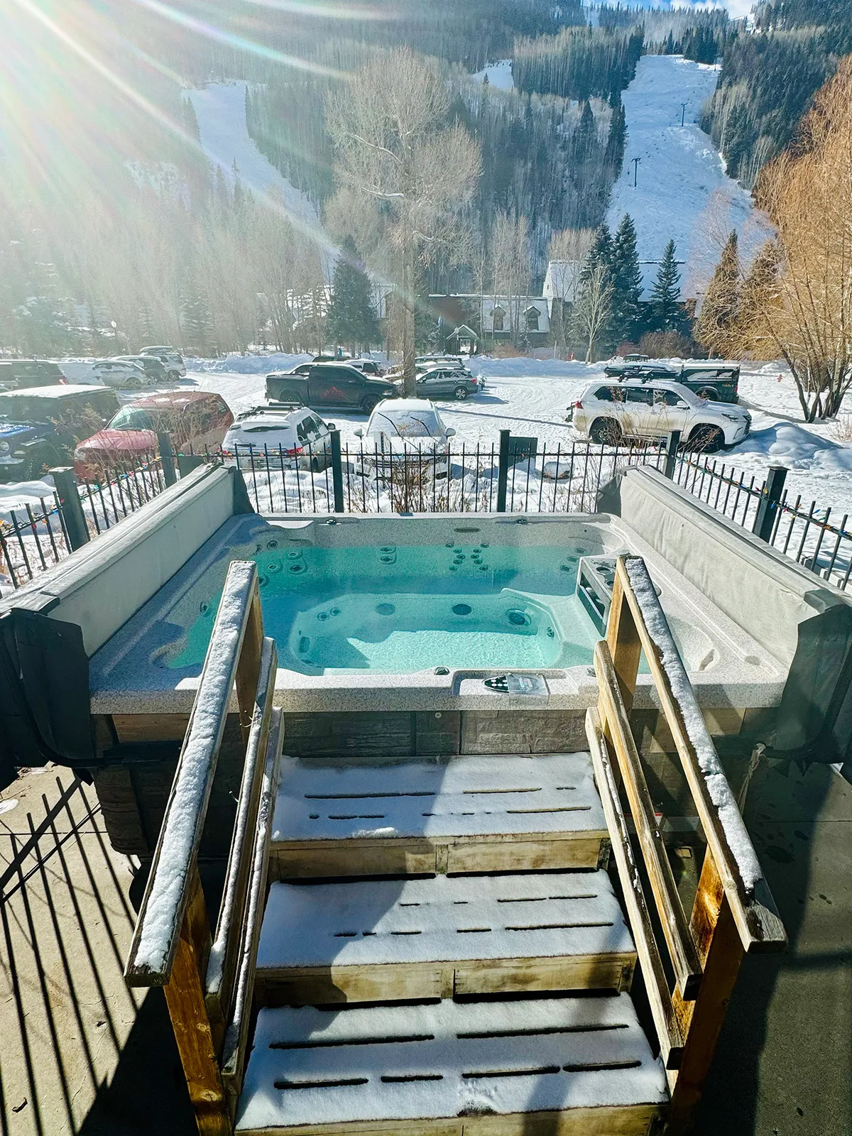 An exterior view of a cozy hot tub surrounded by snow, with mountains rising in the distance. The fenced area and snow-covered steps evoke a retreat-like atmosphere, perfect for unwinding after a day of adventure.
