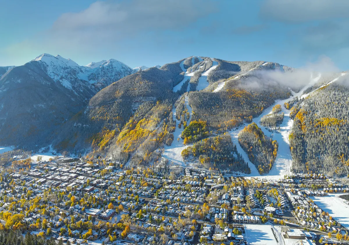 A sweeping exterior view of Telluride with snow-capped mountains rising in the background. The photo captures the vibrant town nestled below, with autumn foliage and ski slopes visible, suggesting a seamless connection between the community and its alpine surroundings.