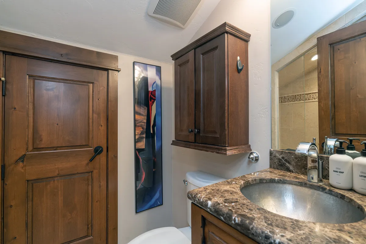 Depicts a warm, inviting bathroom with a wooden door, a granite countertop sink, wall-mounted cabinetry for storage, and colorful artwork adding a personal touch.