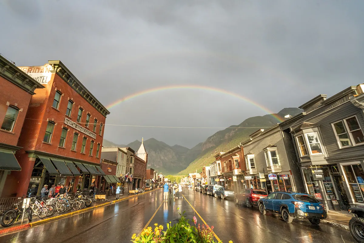 Telluride’s historic main street is alive with people strolling, colorful historic buildings, and a rainbow arching over a damp, sunlit street. The vibrant atmosphere suggests a lively, walkable neighborhood with unique shops and eateries.