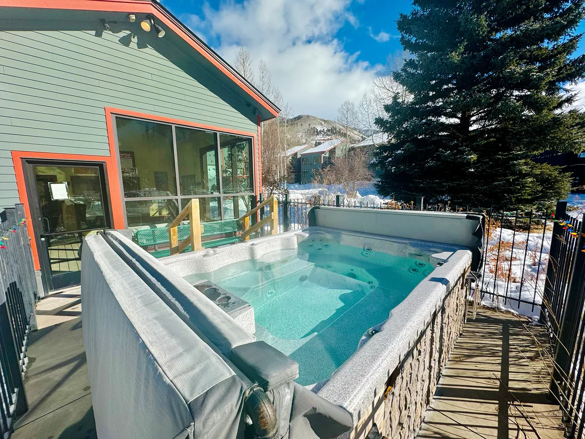 An inviting outdoor hot tub sits on a spacious deck, surrounded by a stunning mountain backdrop. The house exterior features natural wood, and the deck is set up for relaxation and small gatherings. The sun is low in the sky, hinting at a tranquil sunset.