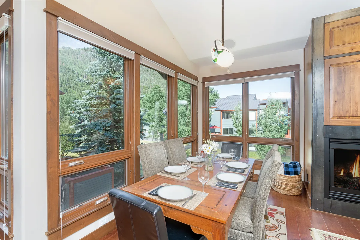 The image highlights a sunlit dining area with a large table set near windows that offer sweeping views of the surrounding landscape. The fireplace adds a cozy touch, while the room’s atmosphere suggests intimate family dinners, story-sharing, and relaxation against a mountain backdrop.