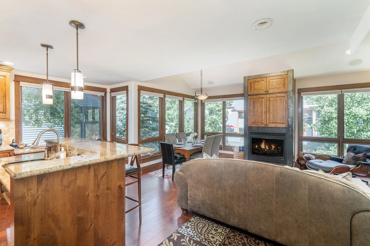 A stylish open-plan living area seamlessly connects the kitchen, dining, and living spaces. The kitchen island, fireplace, and large windows are focal points, creating an inviting space for cooking, dining, entertaining, and relaxing by the fire. The room is illuminated by natural light, enhancing the sense of openness and comfort.