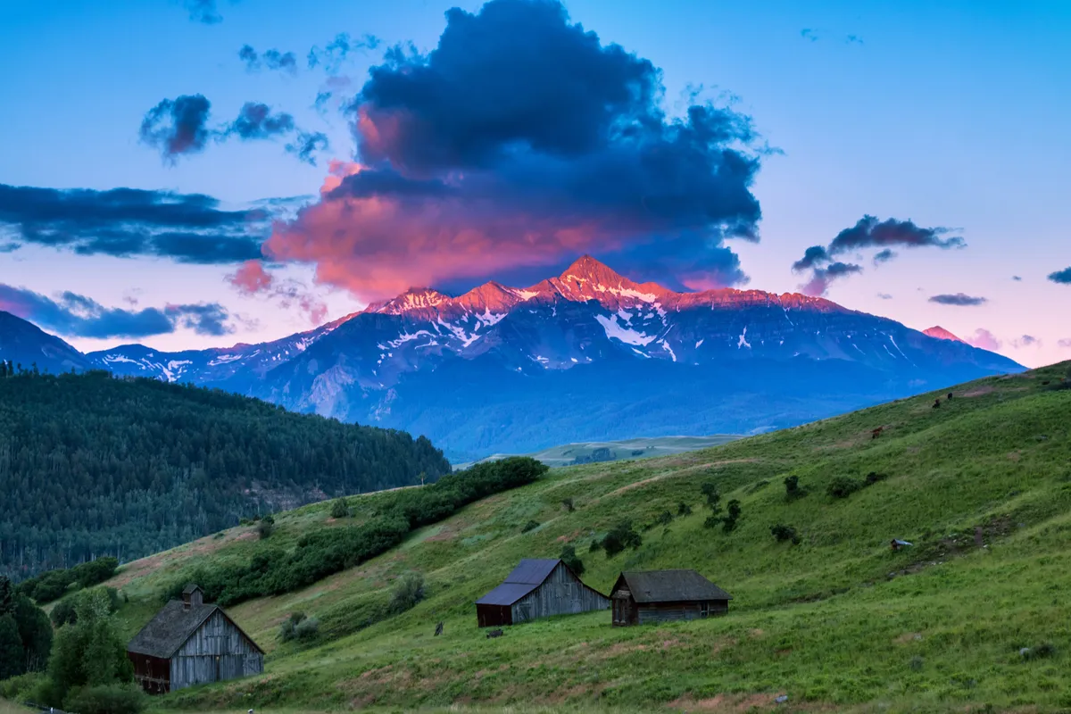 A wide, tranquil exterior shot of lush green hills rolling into majestic mountains, with wooden cabins dotting the landscape. Soft sunset hues paint the clouds, suggesting a peaceful and inspiring natural environment ideal for hikes, photography, or quiet reflection.