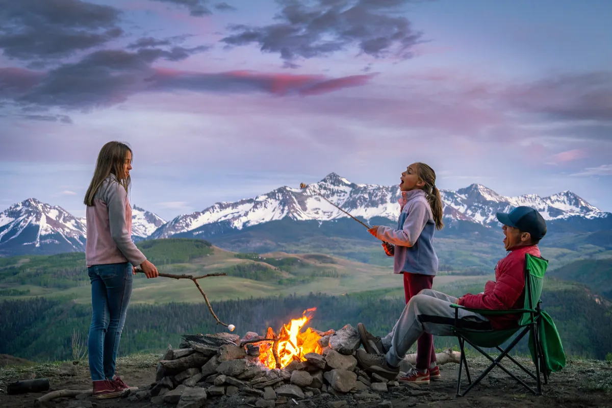 The image shows a group of children and an adult gathered around a campfire, roasting marshmallows in a scenic outdoor area with a mountain backdrop. The setting is playful and serene, emphasizing natural surroundings and family bonding.