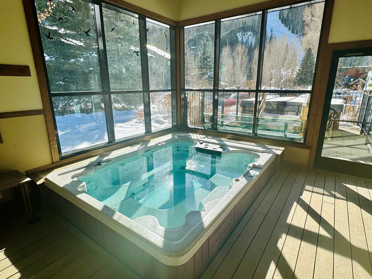 A cozy interior view featuring a modern hot tub set beside large windows, with wooden flooring and abundant natural light flooding the space. It evokes relaxation and comfort—perfect for unwinding after working or adventuring outside.