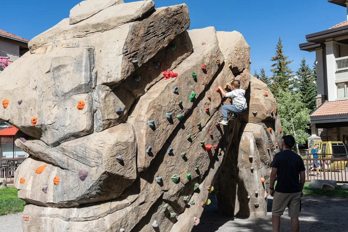 A child climbs a dedicated rock structure outdoors, while an adult supervises nearby. The scene is filled with greenery and open space, suggesting safe and adventurous play for children.