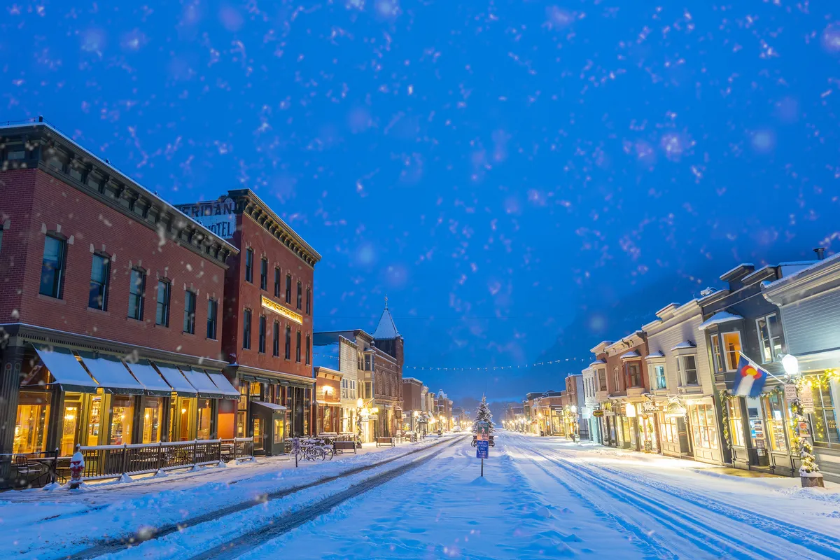 A vibrant exterior street scene with snow-covered buildings, festively decorated shopfronts, and a Christmas tree glowing in the distance. The setting is inviting, with illuminated windows and people strolling, evoking convenience, warmth, and small-town charm.