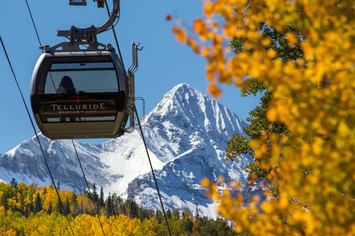 A gondola glides above a colorful landscape of autumn foliage and snow-capped mountains, with the iconic Telluride peaks in the background. The image captures the sense of adventure and accessibility to both hiking and skiing that the area offers.